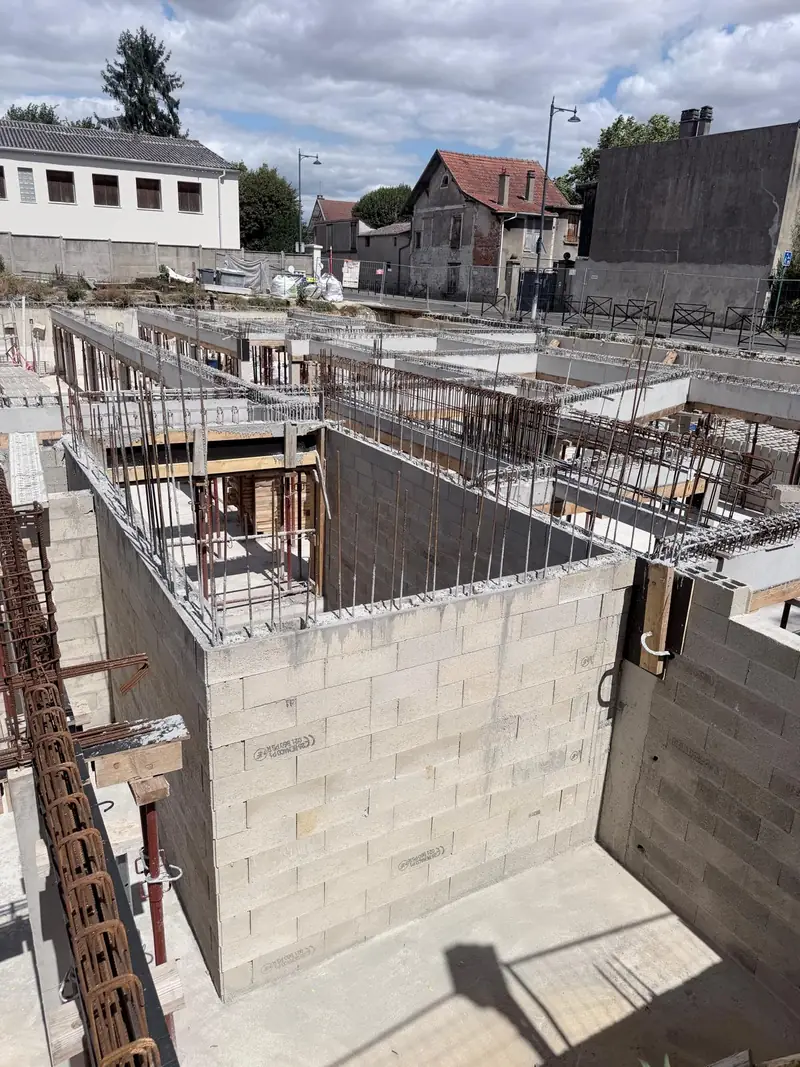 Chantier d’immeuble vu d’en haut : murs en blocs béton, poutres coffrées en bois, étais métalliques rouges, maisons voisines en arrière-plan.