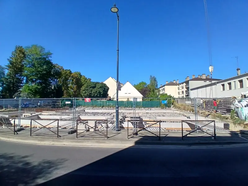 Chantier palissadé vu depuis la rue, ciel bleu, maisons et immeubles voisins.