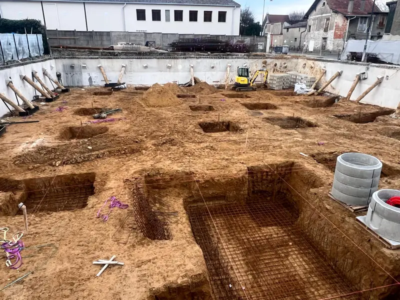 Vue large de la fouille avec tranchées et puits de fondation, parois blanches maintenues par des butons en bois.