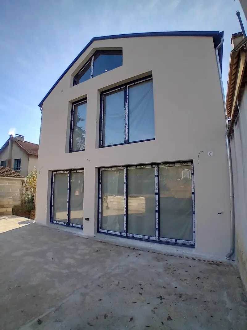 Façade-pignon contemporaine enduite gris clair, rives sombres, grandes baies vitrées noires au rez-de-chaussée, deux fenêtres à l’étage et une baie trapézoïdale sous le toit.