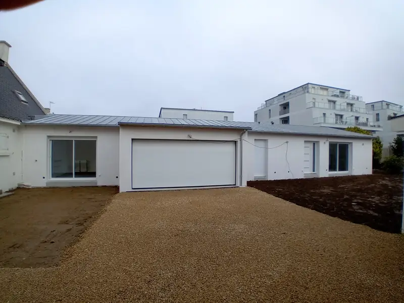 Extension de plain-pied enduite de blanc avec grande porte de garage sectionnelle blanche, baies coulissantes et couverture métallique grise à joints debout, cour gravillonnée au premier plan, résidences récentes en arrière-plan.