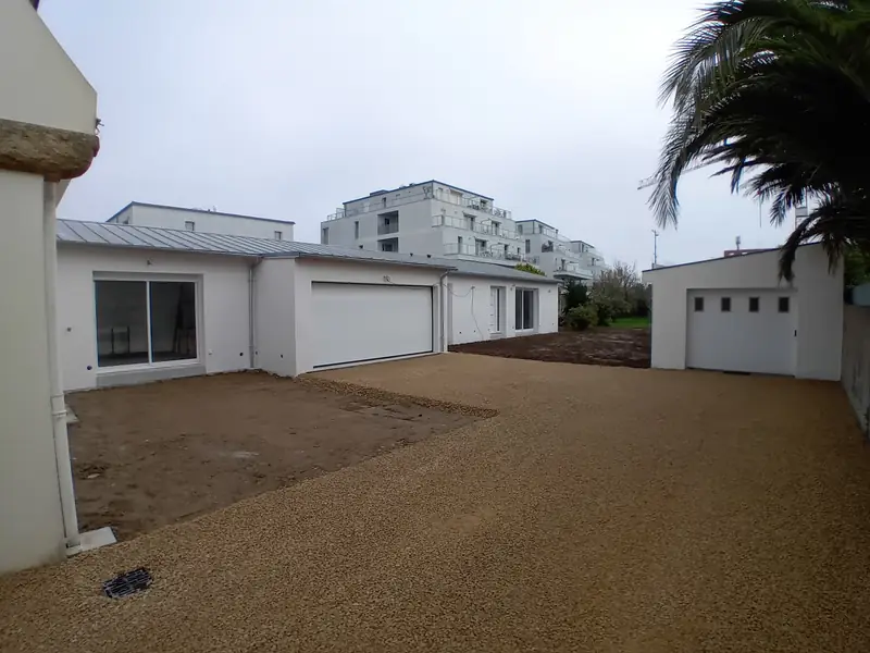 Cour gravillonnée bordée par l’extension blanche à porte de garage et par un garage indépendant en fond de parcelle, palmes au premier plan.
