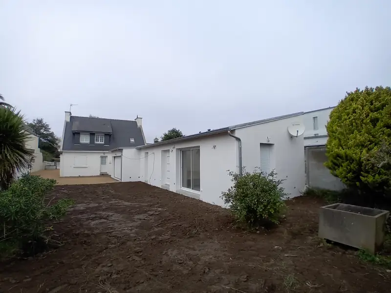 Long volume de plain-pied enduit de blanc, baie coulissante et fenêtres, maison à toit d’ardoise et lucarnes au fond, jardin en terre fraîchement nivelée.