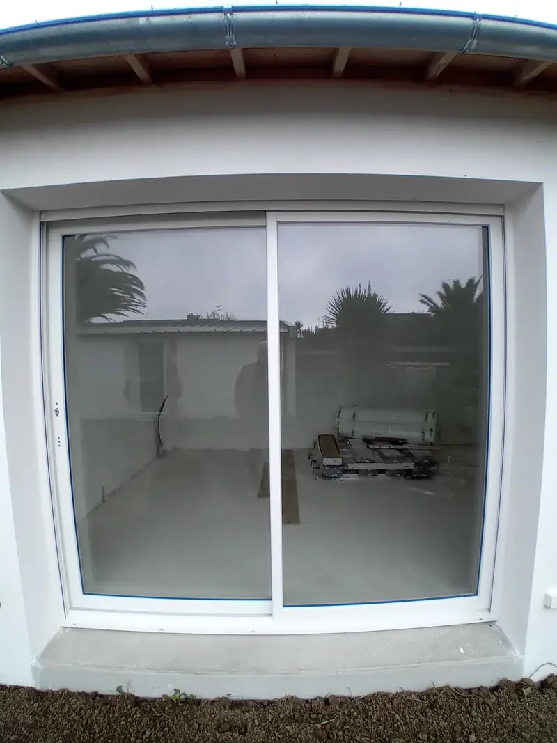 Baie coulissante blanche à deux vantaux dans une façade enduite blanche, seuil en béton, bord de toiture et gouttière au-dessus.