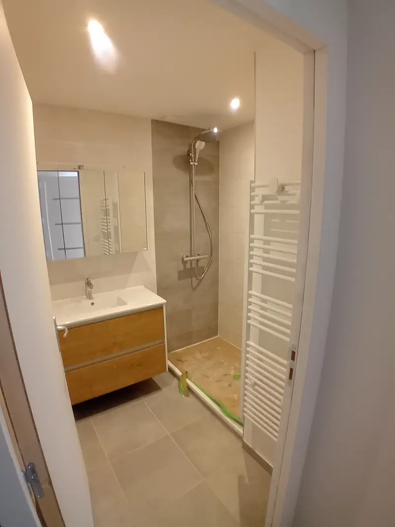 Salle d’eau carrelée gris clair avec douche à l’italienne, colonne de douche, meuble vasque en bois clair, miroir et sèche-serviettes blanc.