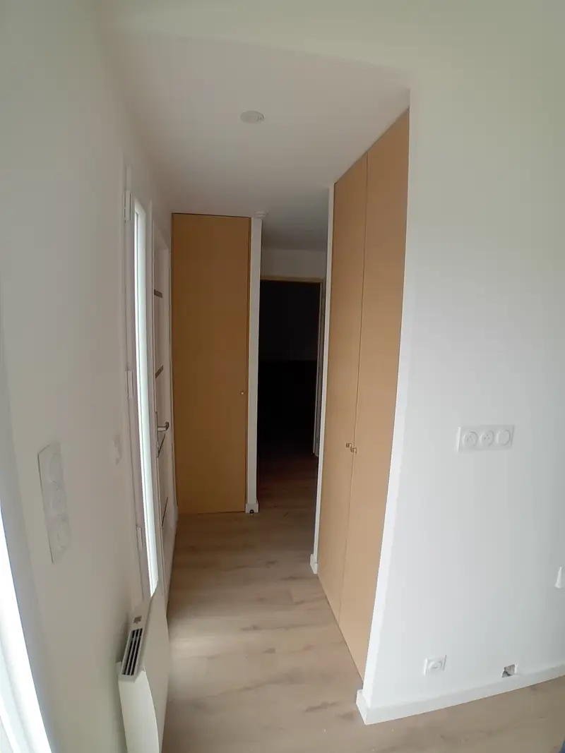 Couloir blanc au sol en lames de bois, placards à portes en bois clair, radiateur électrique et prises murales.