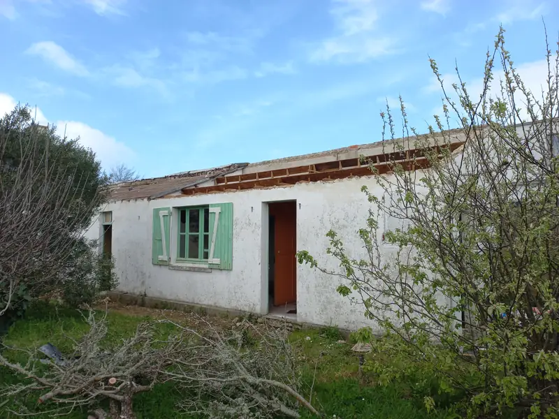 Annexe enduite de blanc avec fenêtre à volets verts et porte orange, charpente à nu, arbustes et branches coupées au premier plan.