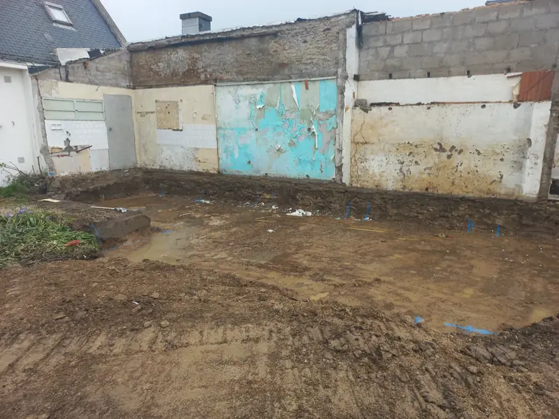 Terrain décapé et détrempé devant un mur ancien couvert de faïence blanche, de peinture turquoise écaillée et d’enduits jaunes.