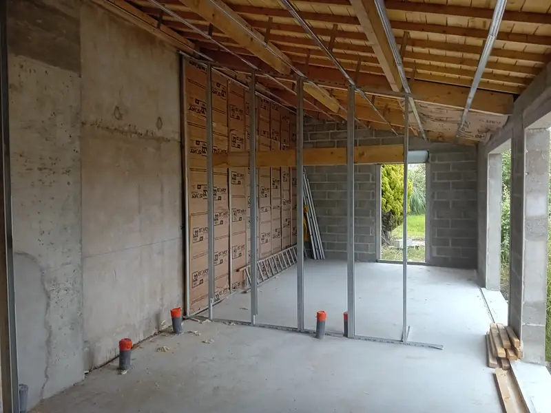 Pièce brute avec mur en béton, montants métalliques de cloison et rouleaux d’isolant en laine minérale, plafond à solives bois.