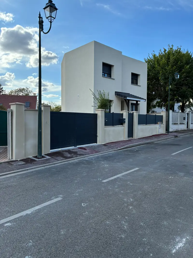 Maison cubique blanche à un étage, deux volumes décalés, menuiseries anthracite, clôture à piliers enduits et portail anthracite, ciel bleu.