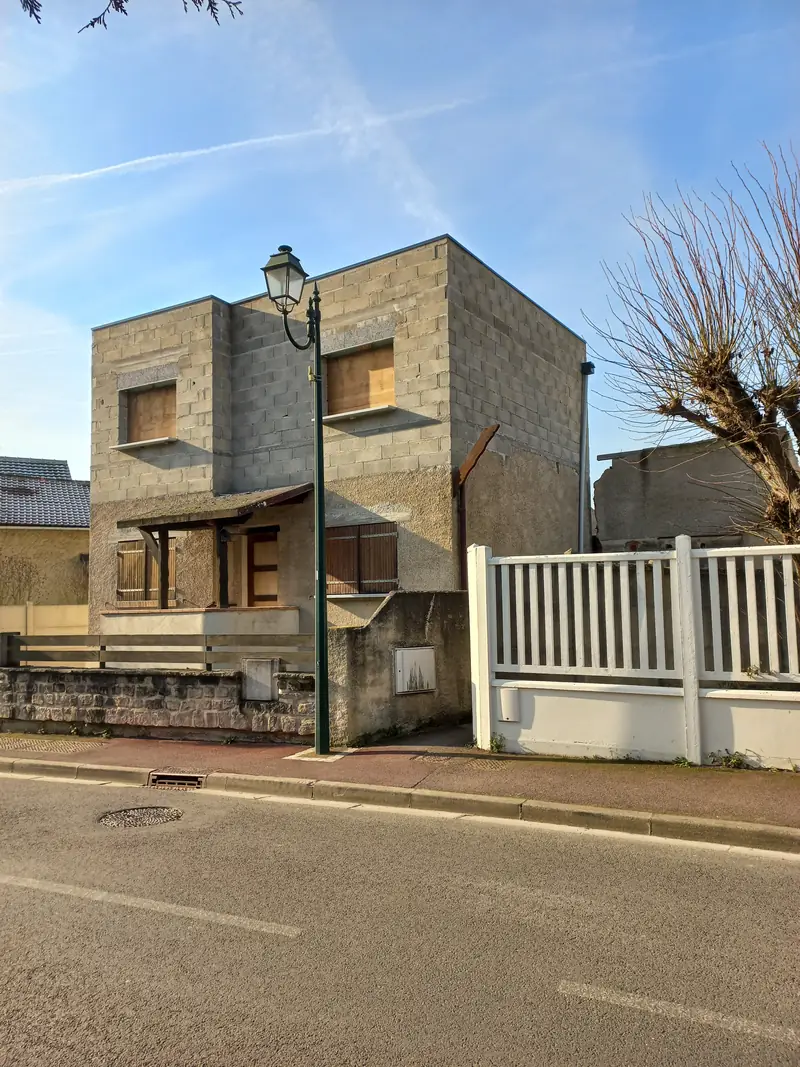 Maison cubique en parpaings bruts non enduits, fenêtres fermées par des panneaux de bois, clôture blanche.