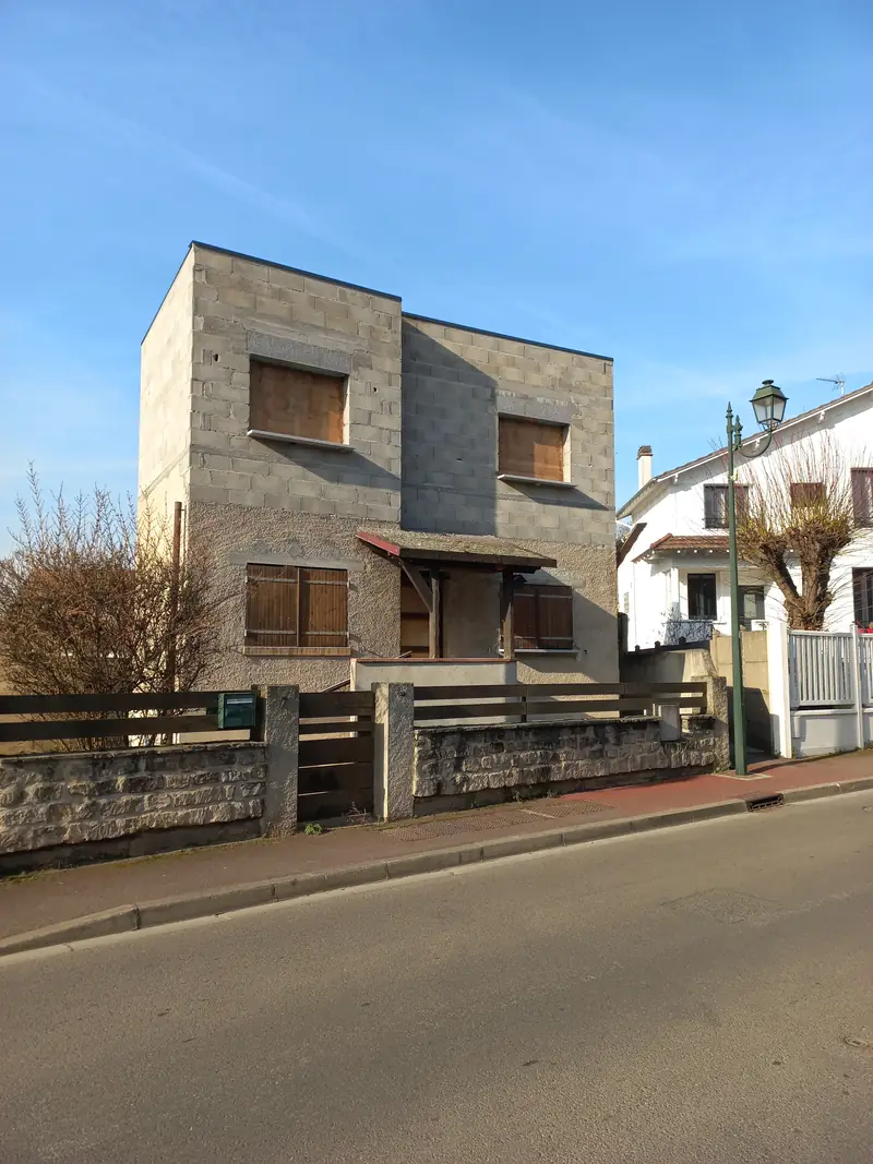 Maison en blocs béton bruts vue de face, auvent en bois, muret de pierre et clôture bois.