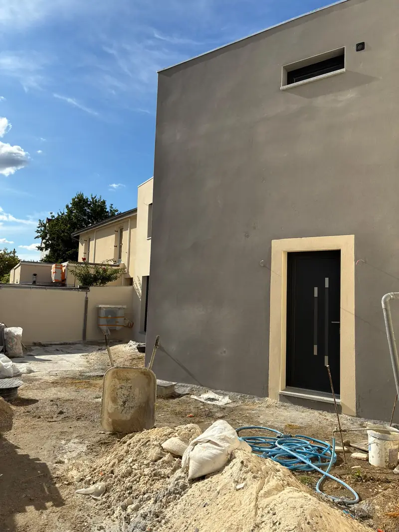 Façade gris foncé avec porte noire encadrée de clair, petite fenêtre haute, jardin en travaux.