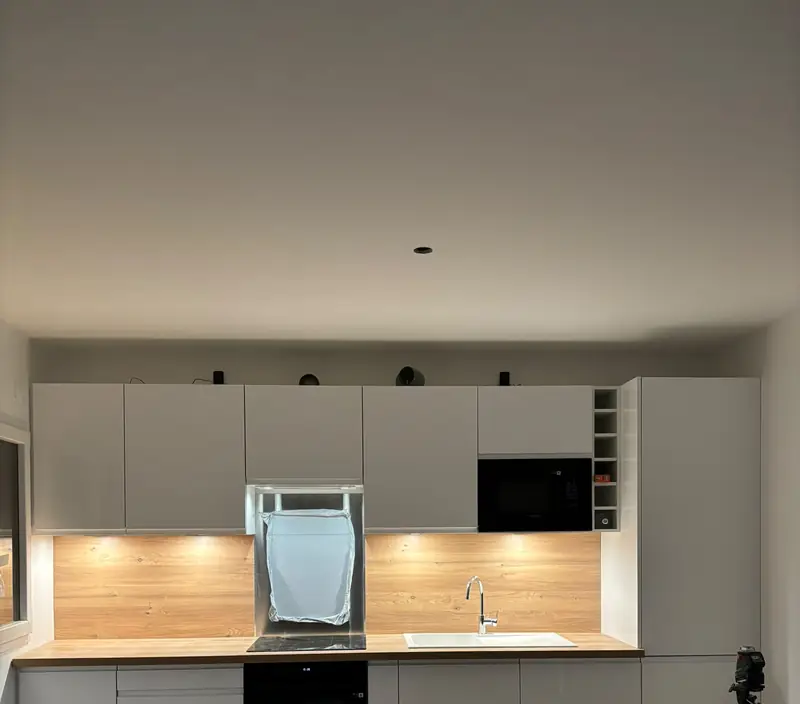 Cuisine blanche linéaire avec plan de travail et crédence en bois éclairés par des réglettes.