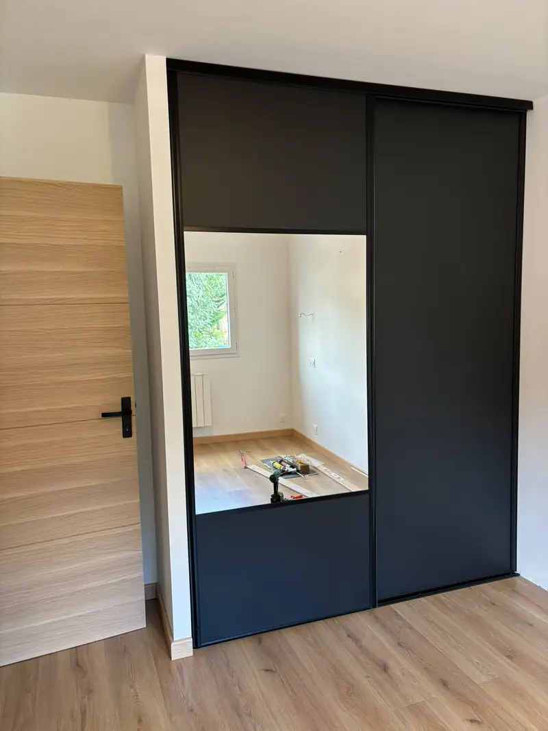 Chambre avec placard à portes coulissantes noires dont une miroir, sol stratifié chêne.