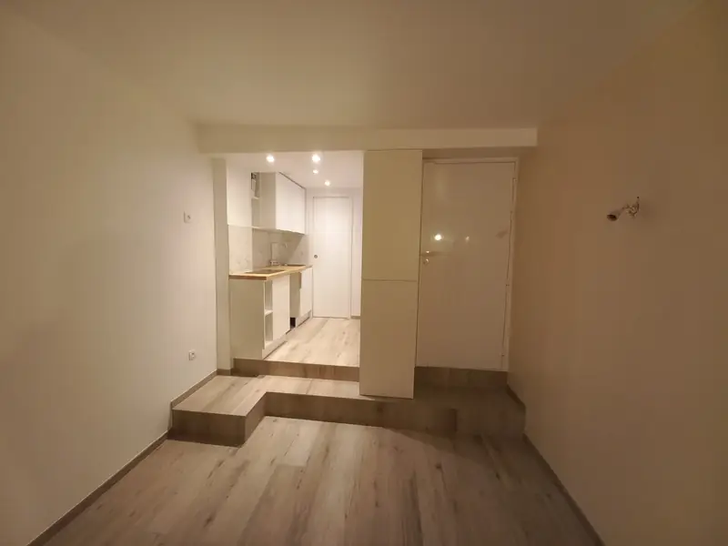 Appartement en longueur : estrade de deux marches menant à une cuisine blanche, porte de salle d’eau, sol stratifié clair.