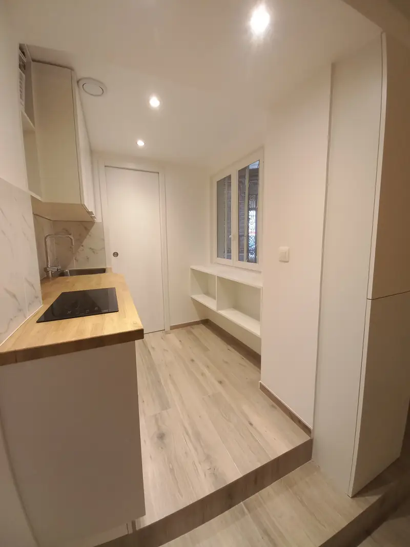 Cuisine étroite sur estrade, plan de travail bois et rangements blancs ouverts sous la fenêtre.