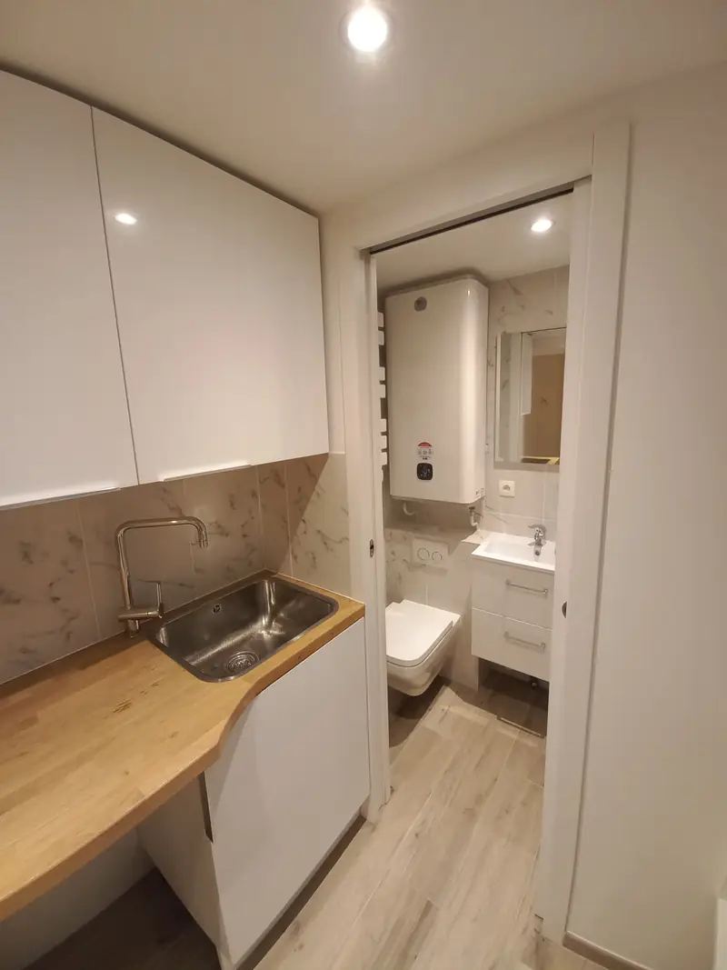 Évier de cuisine au premier plan, porte coulissante ouverte sur une salle d’eau avec WC suspendu et chauffe-eau mural.