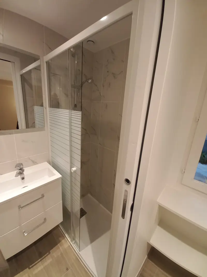Salle d’eau avec douche à paroi vitrée, meuble vasque blanc et faïence effet marbre.