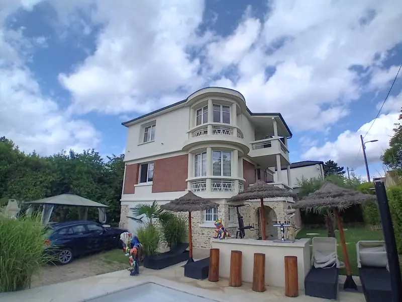 Façade sur jardin de la villa surélevée, piscine au premier plan, ciel nuageux.