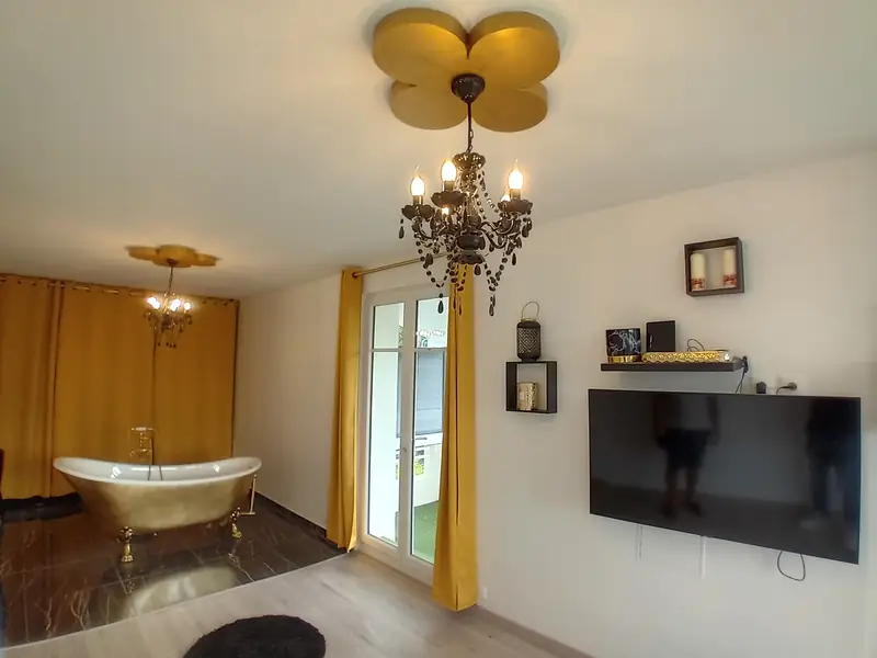 Grande chambre lumineuse avec baignoire îlot dorée sur pieds, lustre, rideaux moutarde et porte-fenêtre.