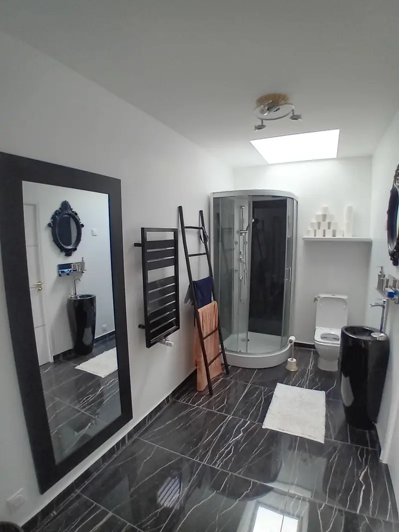 Salle d’eau blanche avec puits de lumière au plafond, carrelage noir veiné, cabine de douche et sèche-serviettes noir.